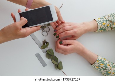 Nail artist taling photo of her work with mobile phone, only hands visible. Green fingernails design, nail files, manicure scissors and eucalyptus branch on white table. Beauty concept.