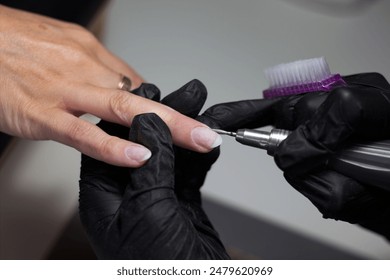 A nail technician, wearing black gloves, uses an electric nail drill on a client’s nail for precise detailing. The client’s hand is comfortably positioned, highlighting the meticulous care in the nail - Powered by Shutterstock