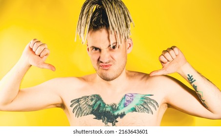 Naked Handsome Young Man 30s With Tattoo Pointing Gesture Of Likes On Himself. Attractive Confident Guy With Blonde Dreadlocks Showing Thumbs Up On Himself In Studio On Isolated Yellow Background