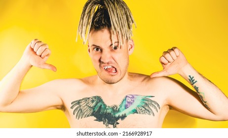 Naked Handsome Young Man 30s With Tattoo Pointing Gesture Of Likes On Himself. Attractive Confident Guy With Blonde Dreadlocks Showing Thumbs Up On Himself In Studio On Isolated Yellow Background