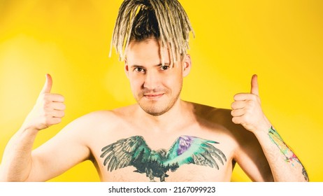 Naked Handsome Young Man 30s With Tattoo On Chest Showing Gesture Of Like. Attractive Smiling Guy With Blonde Dreadlocks Showing Thumbs Up, Looking At Camera In Studio On Yellow Background