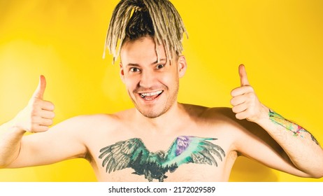 Naked Handsome Young Man 30s With Tattoo On Chest Showing Gesture Of Like. Attractive Smiling Guy With Blonde Dreadlocks Showing Thumbs Up, Looking At Camera In Studio On Yellow Background