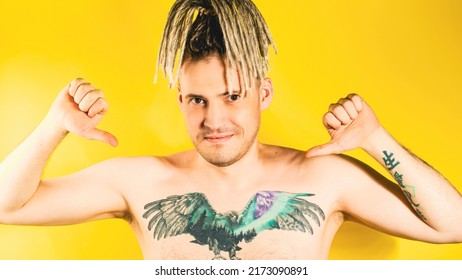 Naked Handsome Young Man 30s With Tattoo Pointing Gesture Of Likes On Himself. Attractive Confident Guy With Blonde Dreadlocks Showing Thumbs Up On Himself In Studio On Isolated Yellow Background