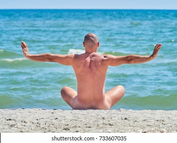 A naked man is sitting on an empty beach and meditating.