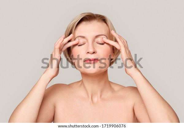Naked\
mature woman standing isolated on grey background beauty concept\
close-up applying eye cream on eyelids\
pensive