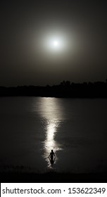 Naked Woman Silhouette Entering Lake Water In The Reflection Of The Moonlight. Full Moon In The Sky And Water Trail Reflection In The Water. Long Exposure.