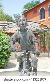 NAKHON RATCHASIMA, THAILAND - April 16, 2016 : Sculpture Of School Boy (or Newspaper Boy) Riding Bicycle In The Garden, Abstract Blur Of Italian Style Architectural Building And Park, Palio Khao Yai. 