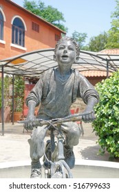 NAKHON RATCHASIMA, THAILAND - April 16, 2016 : Sculpture Of School Boy (or Newspaper Boy) Riding Bicycle In The Garden, Abstract Blur Of Italian Style Architectural Building And Park, Palio Khao Yai.