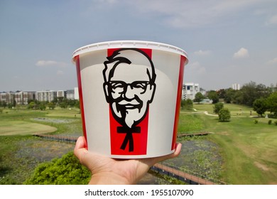 Nakorn Pathom, April 8 2021: KFC Bucket In The Hand Of South East Asian Male Left Hand, Raising The KFC Family Bucket Set For Six People With Colonel Sanders Face Logo Up With Sky As A Background.