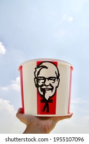 Nakorn Pathom, April 8 2021: KFC Bucket in the hand of South East Asian male left hand, raising the KFC Family bucket set for six people with Colonel Sanders face logo up with sky as a background.