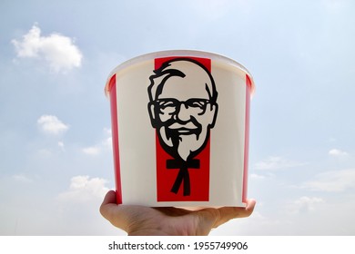 Nakorn Pathom, April 8 2021: KFC Bucket In The Hand Of South East Asian Male Left Hand, Raising The KFC Family Bucket Set For Six People With Colonel Sanders Face Logo Up With Sky As A Background.
