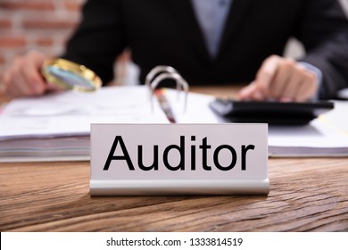 Nameplate With Auditor Title Kept On Desk In Front Of Businessman Examining The Invoices