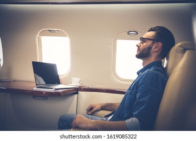 A nap while flying. A side-view photo of a young man, who is sitting near the desk with a laptop on it, in his window seat on the board of the plane, and taking a nap.