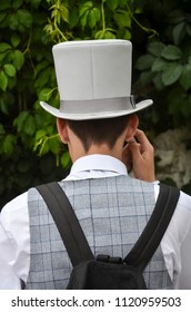 the nape of a young man in a gray cylinder and a checkered waistcoat with a backpack on his back against a background of green foliage. Man with hat from behind.