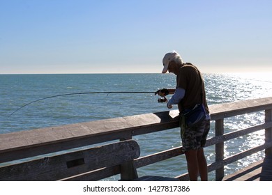 Naples, FL/USA: Feb 28, 2019 - Fisherman With Grey Hair On Naples Pier In Florida At Dusk Handles His Fishing Rod In Cloudless Blue Sky As Setting Sun Sparkles On Gulf Of Mexico.
