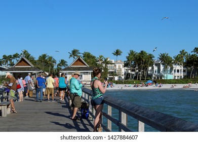 
Naples, FL/USA: Feb 28, 2019 - Visitors And Fishermen Stand On The Pier, Known For Exquisite Sunsets And Fishing. Sandy Beach, Swimmers, Sunbathers, Palm Trees And Luxury Housing In Background. 