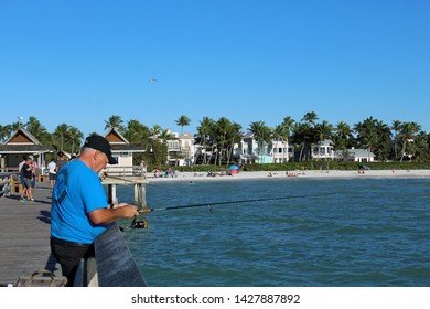 Naples, FL/USA: Feb 28, 2019 - Fisherman Stands On The Pier, Known For Exquisite Sunsets And Fishing. Sandy Beach, Swimmers, Sunbathers, Palm Trees And Luxury Housing In Background.