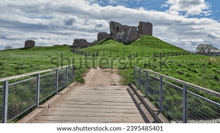 Narrow bridge through green valley leads to the ruins of Duffus Castle, located on grassy hill near Moray, Scotland, under blue cloudy sky