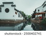 A narrow canal in Suzhou, China runs between two white buildings with traditional Chinese architecture.