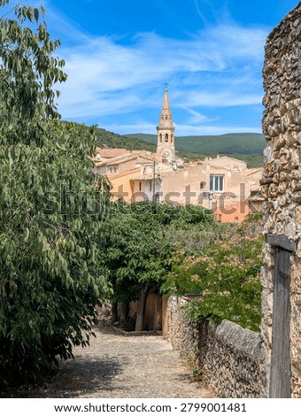 A narrow, cobblestone path lined with natural stone walls winds through the Mediterranean village of St-Saturnin-lès-Apt in Provence, leading to a striking church tower