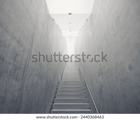 Narrow concrete steps leading up to the light. Empty staircase with handrails on the right side, concrete walls and illuminated satin glass ceiling. Almost monochrome photo, cold tones.