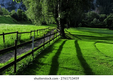A narrow dirt path lined with a rustic wooden fence cuts through a manicured golf course, where sunlight casts long shadows of trees across the vivid green grass. Lush forests and rocky cliffs frame the scene in the background.
