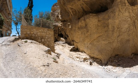 A narrow dirt path winds between a stone wall and a rocky cliff under a clear blue sky.