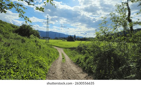 A narrow dirt path winds through lush greenery, leading to open fields under a partly cloudy sky with distant hills and power lines in view. Peaceful and scenic countryside. - Powered by Shutterstock - Get 15% off with code: PIKWIZARD15