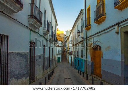 A narrow laneway in a gritty quarter of the medieval town of Cáceres, Extramadura, Spain. The laneway leads up to the Plaza Mayor (main square) under a sunny blue sky.
