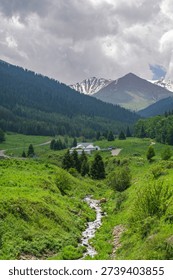 A narrow mountain stream flows through a lush green ravine, leading the eye toward a yurt camp and high snow-capped summits under a heavy, atmospheric sky.