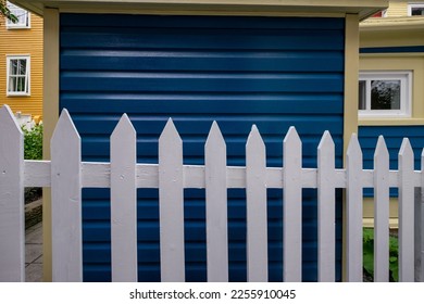 A narrow navy blue wood Cape Cod siding exterior of a vintage building with white trim around the window. There's a country style white wooden picket fence in the foreground with lats and rails. - Powered by Shutterstock