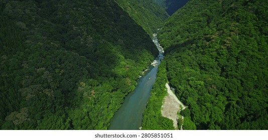 A narrow river winds through a deep valley covered in thick green forest foliage between two large mountains under bright light in this natural landscape view of the wilderness
