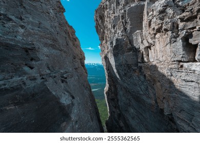 A narrow rocky passage between two large cliffs, revealing a distant view of a green valley and snow-capped mountains under a clear blue sky. - Powered by Shutterstock