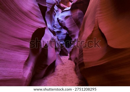 A narrow sandy pathway winds through a slot canyon with smooth, curved sandstone walls. The rock formations display rich colors ranging from deep purple to pink and red with natural layered striations.