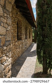 Narrow stone walkway between a rustic stone chapel wall and cypress trees, with sunlight and shadows in a peaceful Mediterranean courtyard.