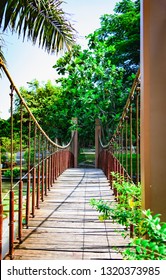 A Narrow Suspended Wooden Bridge Hangs On A Rope Above The Water.  Tropical Park In Vietnam With Green Vegetation