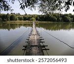 A narrow suspension bridge stretched across a calm river, on the other side of which lush trees grow, creating a wonderful summer atmosphere and inspiring thoughts.