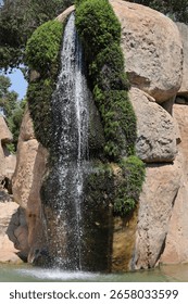 A narrow waterfall flows down between moss-covered rocks, surrounded by large sandstone boulders and lush greenery, creating a peaceful and natural oasis.