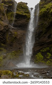 A narrow waterfall flows down a rugged, moss covered cliff into a shallow pool with mist rising, surrounded by rocks and a small stream under an overcast sky.