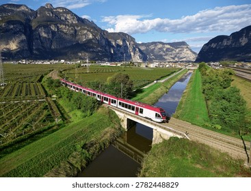 A narrow-gauge electric multiple unit train operating on the Trento–Malè–Mezzana railway, dashing through the beautiful landscape of vineyard fields on a sunny day and a rugged mountain wall dominating the background, in San Michele all'Adige, Trentino, Italy.

The photo shows the line running through the flat vineyard valley near Mezzocorona in the Trentino region. It is crossing a small stone bridge over a canal, set against the dramatic backdrop of towering Limestone cliffs.

The picture captures a wide, flat agricultural valley surrounded by dramatic alpine rock formations under a bright sky.
Massive, steep rocky mountains with vertical gray stone faces border the entire valley background.
The valley floor is filled with neatly organized green fields, patterned with rows of agricultural crops or vineyards.
A long, straight drainage canal with reflective water cuts through the green landscape, framed by grass banks.
