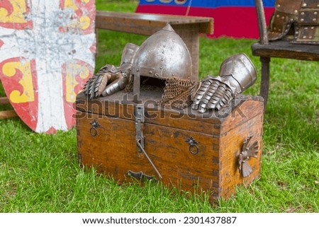 A Nasal helmet (chichak) and a pair of Gauntlets on the top of a wood chest next to a shield the with Byzantine imperial coat of arms of the Palaiologos dynasty.