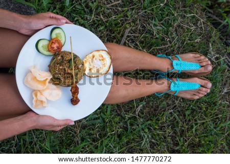 Nasi goreng, traditional Indonesian dish served with prawn crackers and chicken satay on white plate. Closeup woman's hands holding plate with fried rice while sitting on the green grass