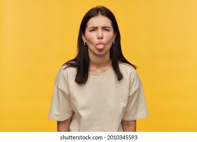 Nasty playful brunette young woman in white tshirt looks naughty and showing tongue over yellow background Looking at camera