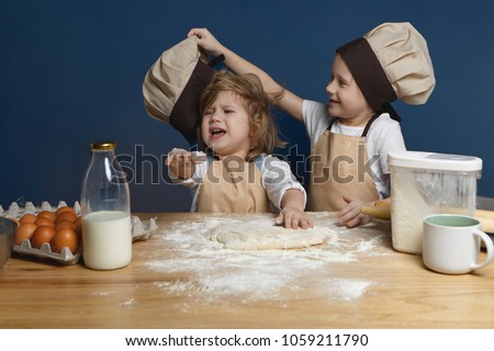 Nasty schoolboy in chef uniform teasing his crying little sister, taking off her cap while making dough together in kitchen, standing at counter with dough, milk, flour and eggs, baking cookies