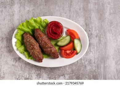 The National Dish Lyulya-kebab With Vegetables And Lettuce Leaves With Red Sauce In A Plate On A Gray Background. Horizontal Orientation, Top View, No Face, Copy Space