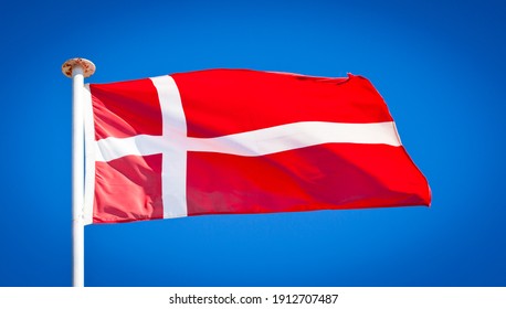 National flag of Denmark. Known as the Dannebrog. Danish flag blowing in strong wind against a pure blue sky. Red with white Scandinavian cross. The flag is the oldest continuously used national flag.