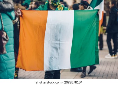 National Flag Of Ireland In Hands Of Unrecognizable Man On Background Of People In City. Saint Patrick Day Holiday