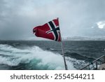 A national flag of Norway flowing in the wind on a fjord tour with a foamy trail left behind the vessel