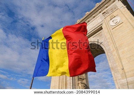 The national flag of Romania winding next to Arch of Triumph landmark building from Bucharest during a sunny day.