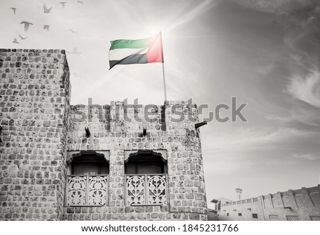 National flag of UAE flying on an old, ancient Arabic house. Black and white, archival photo of Emirati house with color national flag.